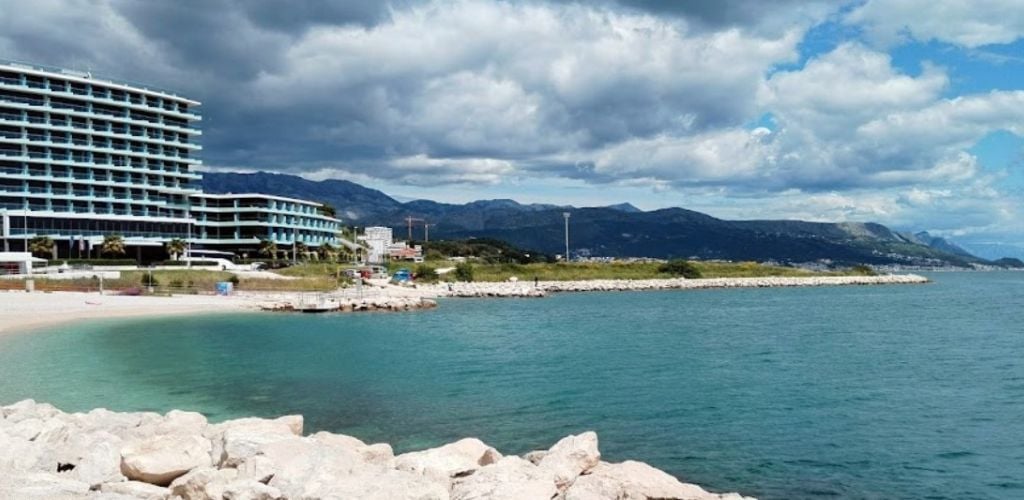 A beach and clean sea water, a small boat at the shore, and a beautiful hotel near the beach. 