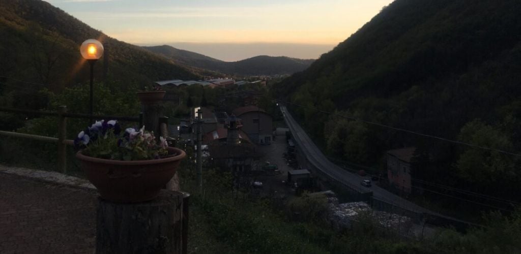 During a sunset, houses and a road in the center between green and a large mountain. 
