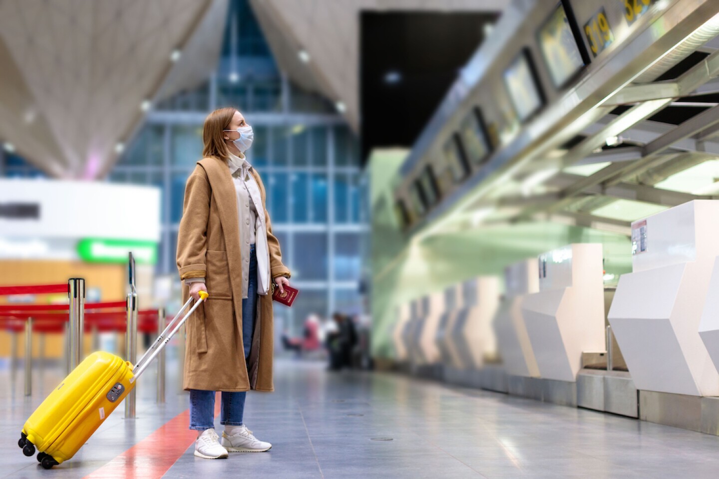 A person in a long brown coat wearing a mask and holding a yellow rolling carry-on suitcase looks at the departures screen at an airport