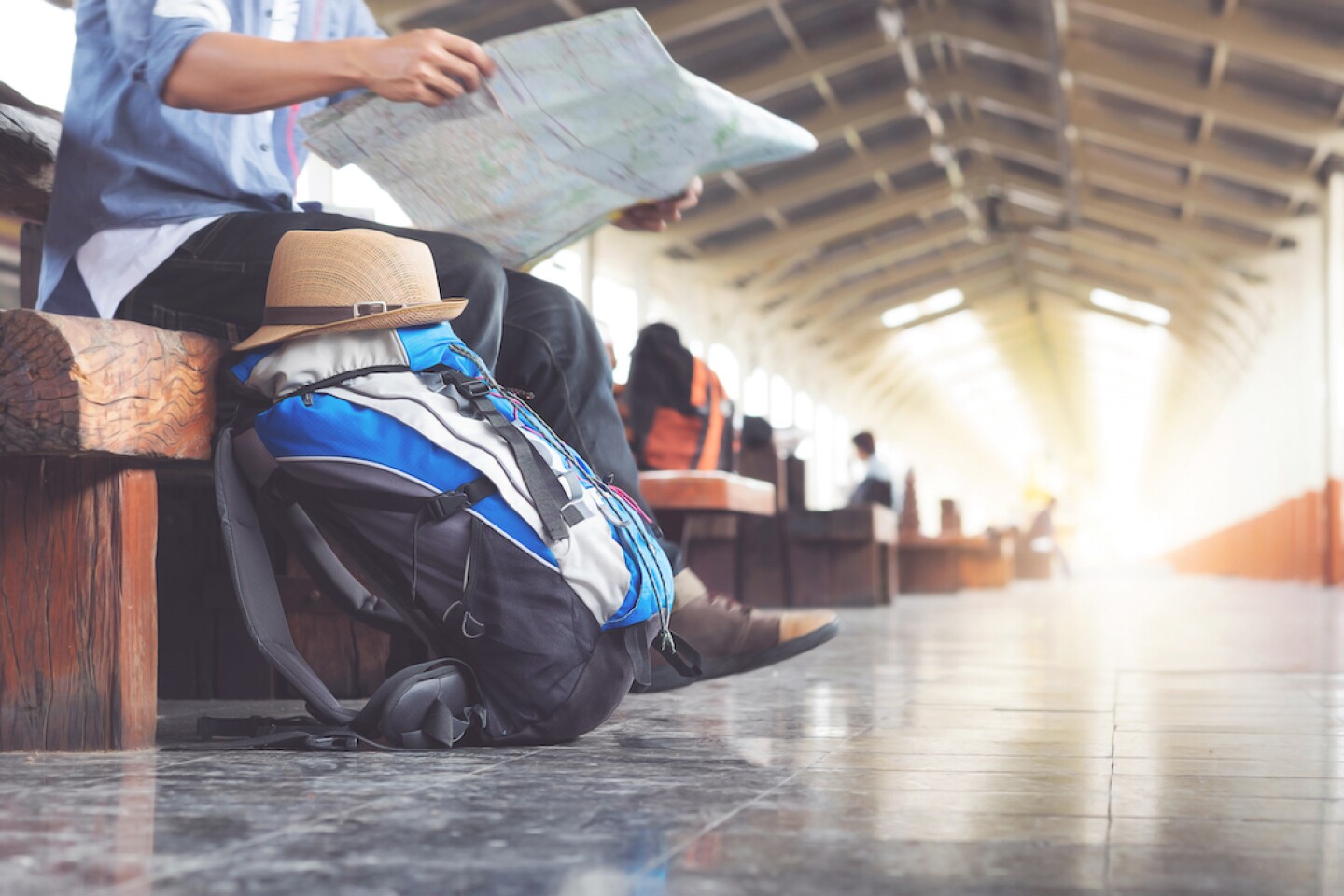 A person looking at a paper map sits on a bench in what seems to be a train station, their backpack on the ground next to them