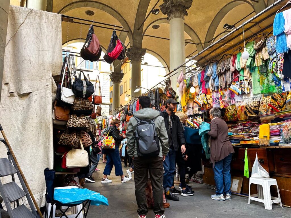 A vibrant street market scene at Mercato del Porcellino in Florence, Italy, featuring a variety of products including leather goods and silk scarves.