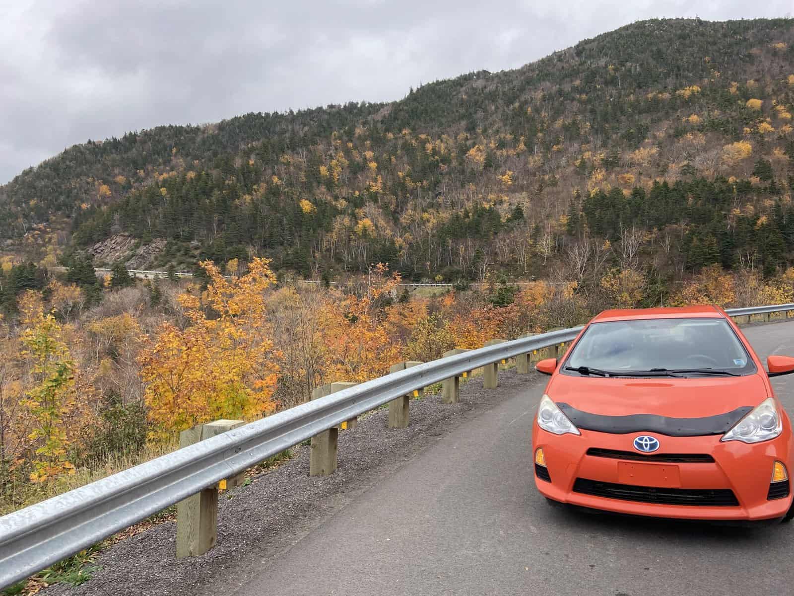 Orange car on the side of the highway with fall foliage behind on a road trip on the Cabot Trail on Cape Breton Island, Nova Scotia; cross Canada road trip