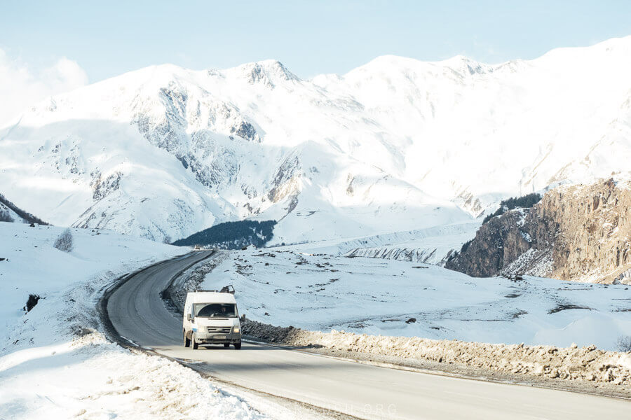 A white van travels down the Georgian Military Highway, a high road covered with snow and surrounded by frosty mountains in Georgia.