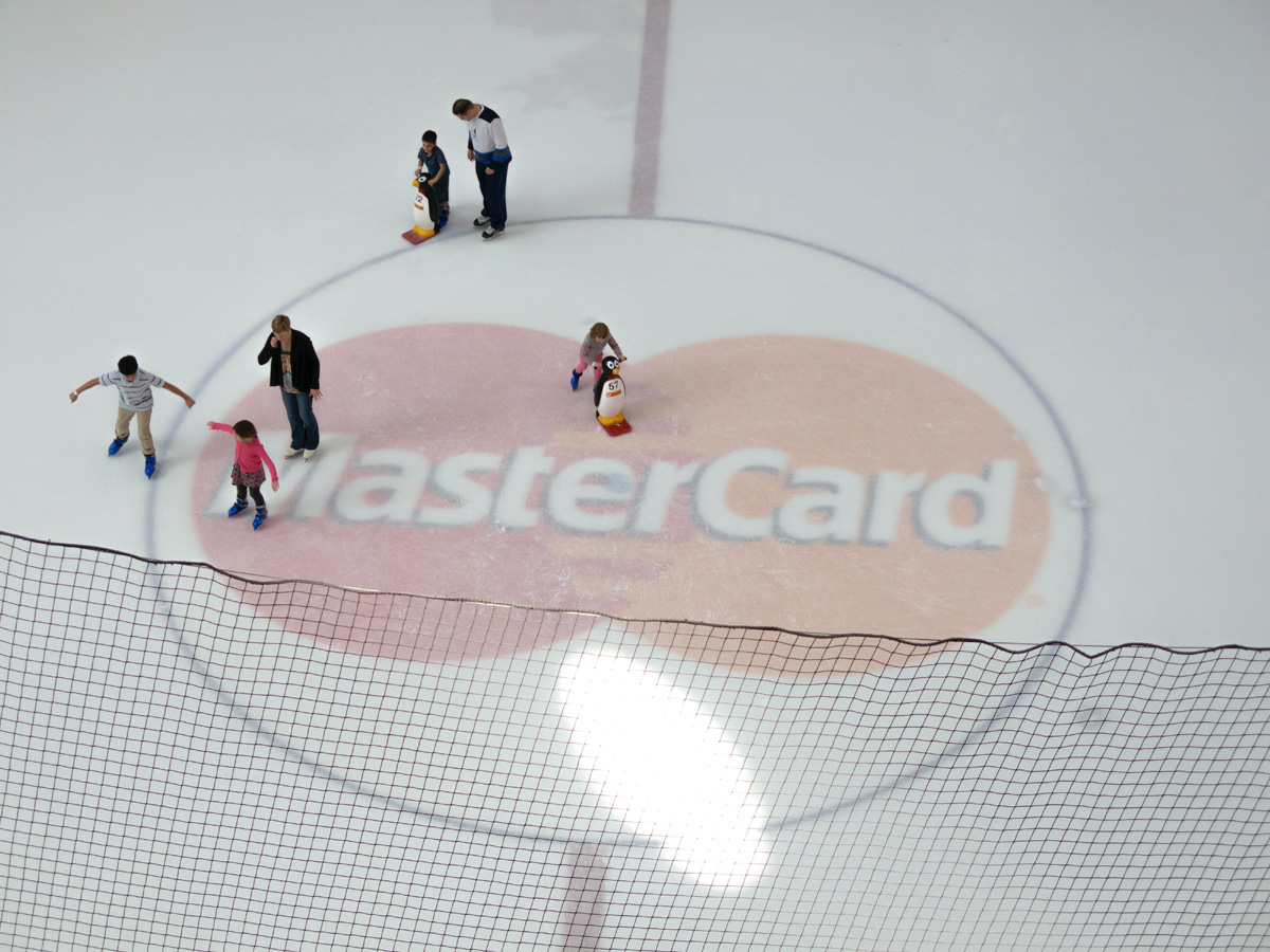 A sweeping white ice skating ring in Dubai with half a dozen people skating across a MasterCard logo in the ice. 