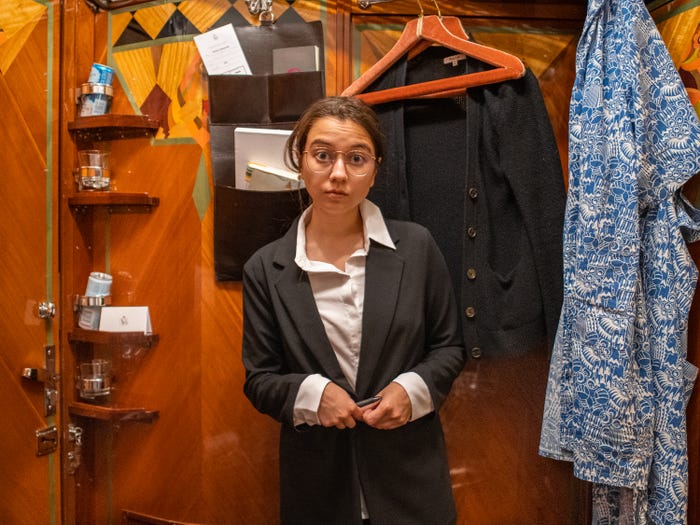 The author stands in front of a red wooden wall with painted details in a black and white suit with more clothes hanging behind her