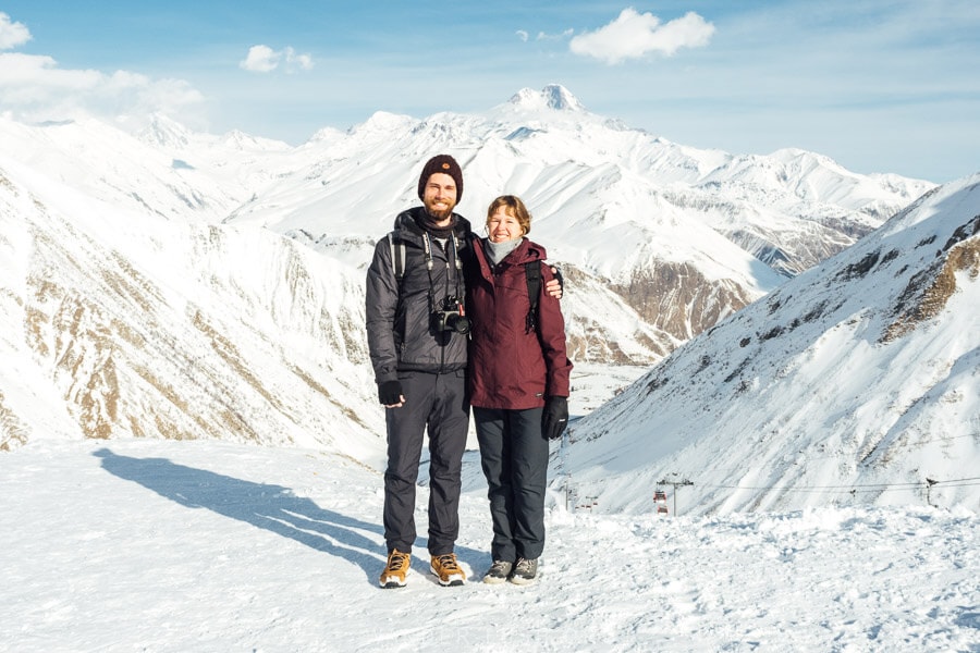 A man and a woman dressed in ski clothes pose for a photo in front of Mount Kazbek in Georgia.