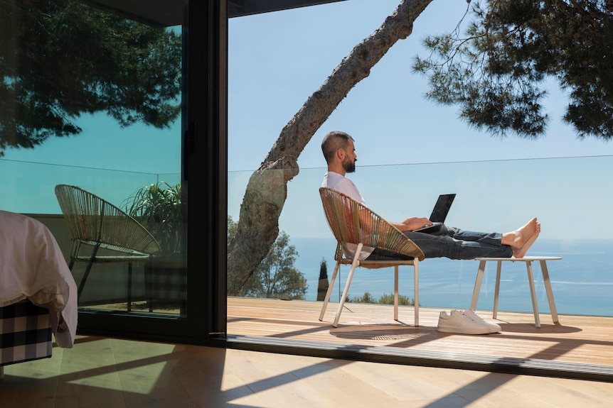 Man sitting on a chair with his legs up with his laptop on a balcony overlooking the ocean