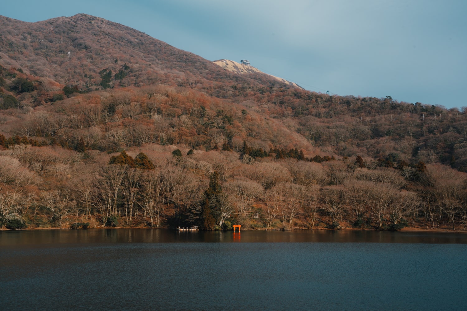 A mini red torii gate in the water with aerial tram station on hill in the background at Lake Ashi, Hakone, Japan.