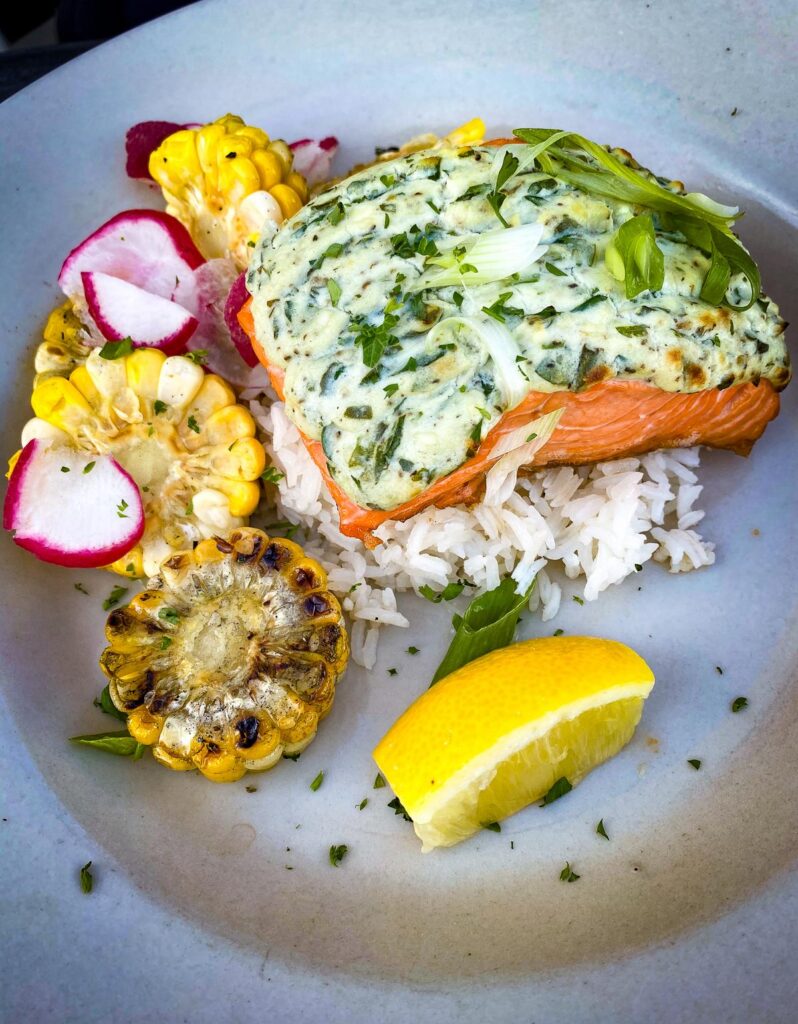 A high-resolution image of an Alaskan salmon dish with herb sauce, grilled corn, radishes, and rice, served at a renowned Fairbanks restaurant.