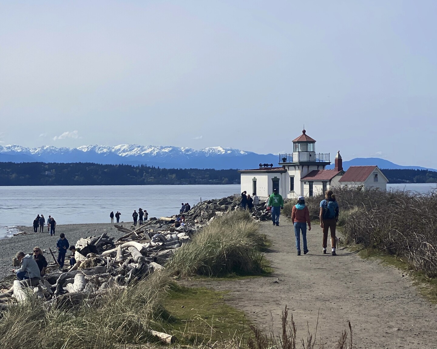 Short white lighthouse with red roof in Discovery Park in Seattle, with water and snowcapped mountains in background and a few people walking on trail in foreground