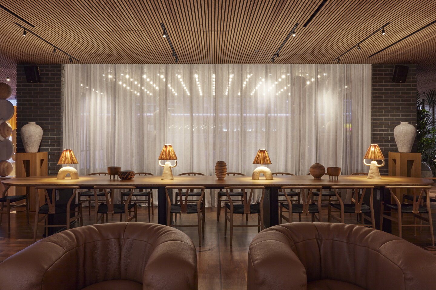 Long table with chairs and topped with small lamps, with two tan curved armchairs in foreground in hotel lobby of One Hundred Shoreditch