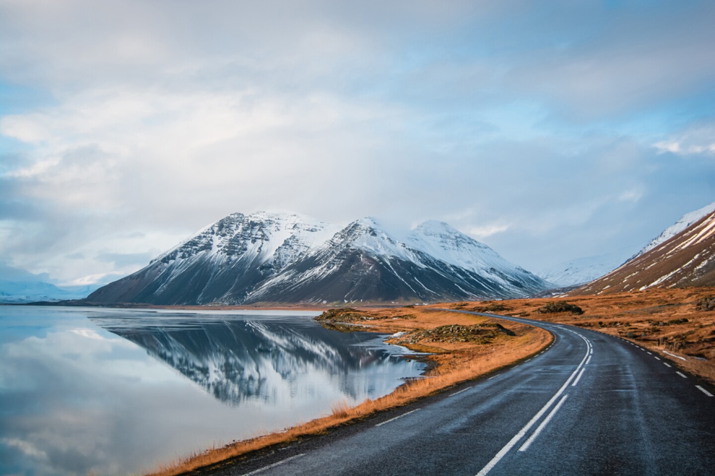 Driver's point of view on Ring Road, Iceland, with empty road leading along coast of lake to volcanic mountains; rocky peaks covered with snow mirrored in lake.