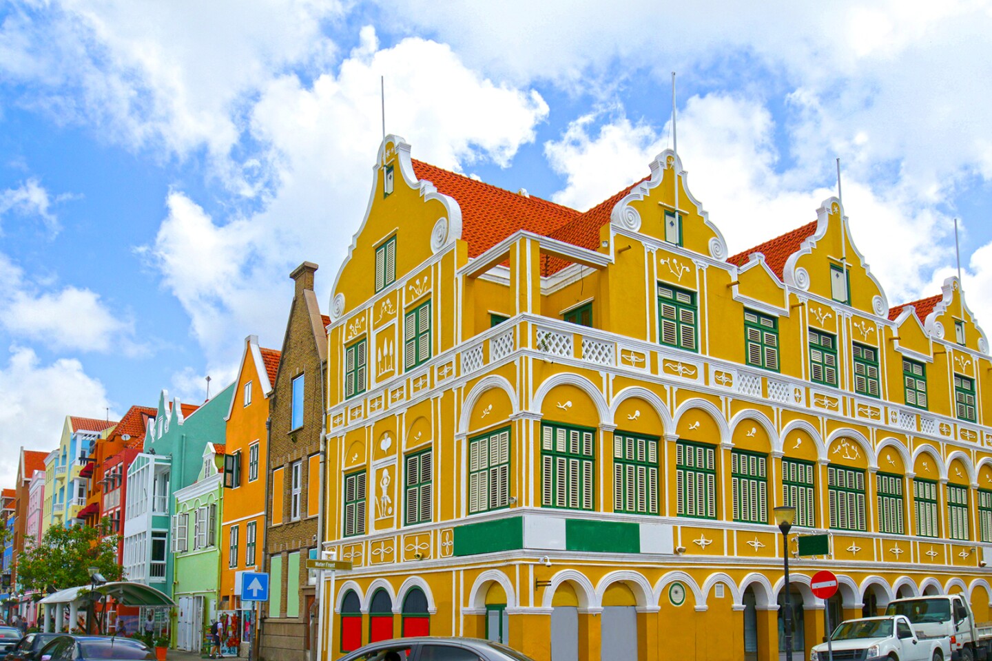 Bright yellow building with a red roof on a street corner, with row of multi-colored historic houses at left