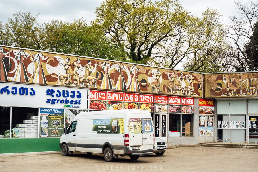 A van parked in front of a row of shops wrapped in an epic mosaic panel in the town of Gurjaani, Georgia.