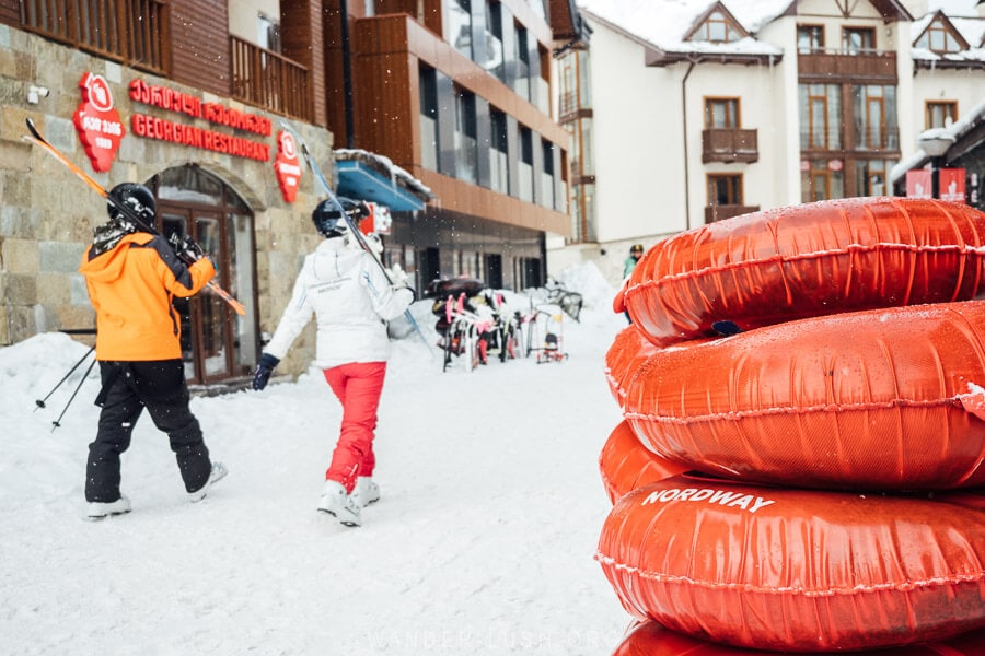 A stack of snow tubes in front of a ski hire shop in New Gudauri, with people walking with skis over their shoulders towards the chalets.