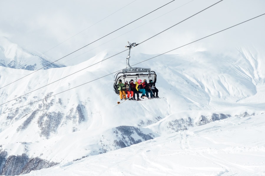 Six friends ride a ski lift in Gudauri, passing through a mountainous valley covered in snow.