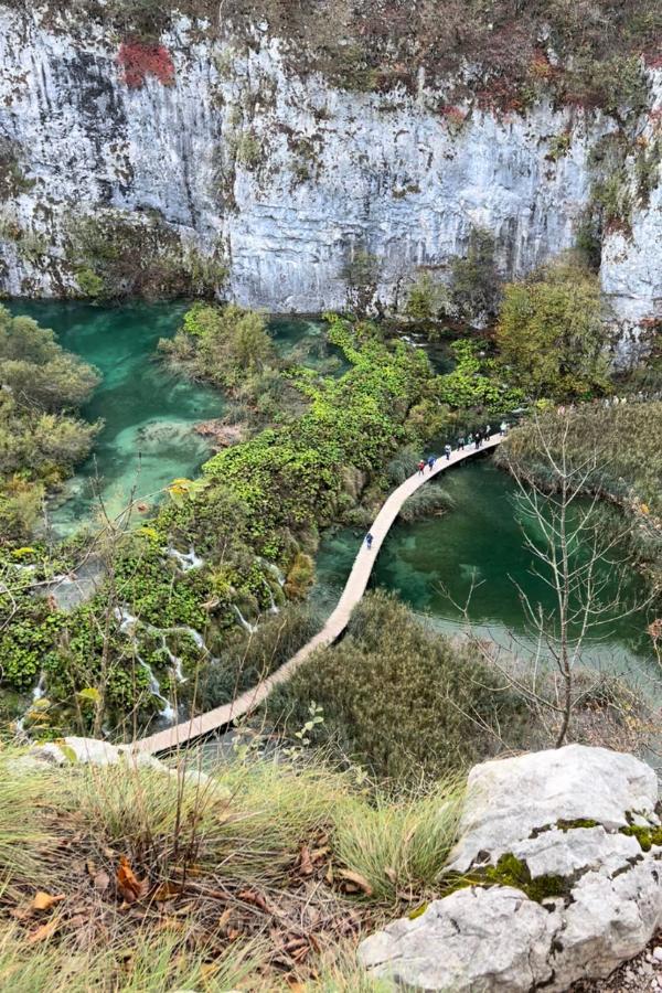 Aerial view of a wooden walkway that crosses over two blue-green lakes in Plitvice National Park in Croatia surrounded by towering cliffs.