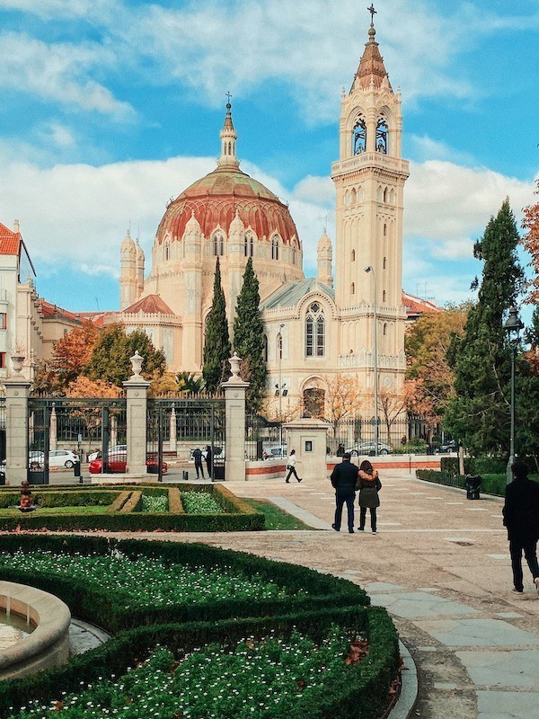 A vibrant view of a cathedral in Madrid with blue skies and fluffy white clouds above. In the foreground are beautifully manicured gardens.