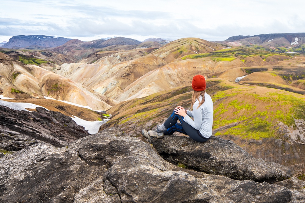 A woman wearing an orange winter hat with her back turned to the camera sitting on large rocks looking out at the hilly landscapes of Iceland.