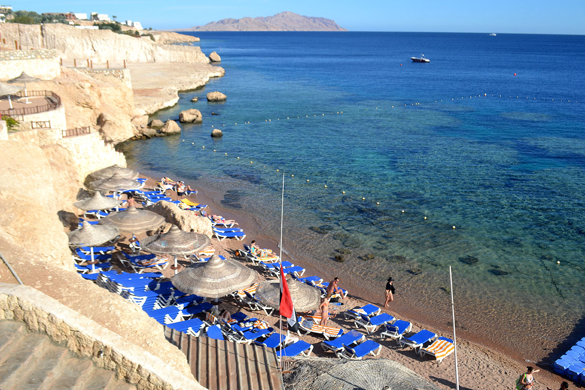 A beach on the Red Sea in Egypt has deep blue waters and lounge chairs lined up alongside some sandy hills. 