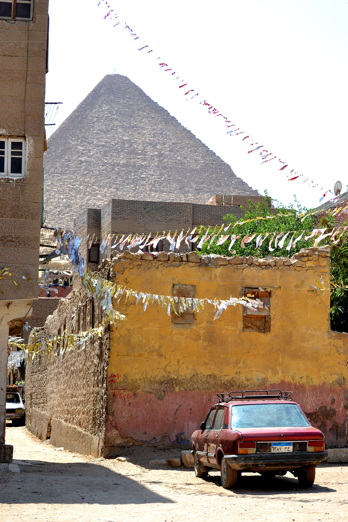 A residential side street in Giza with local homes and a parked car with an enormous pyramid looming in the background. 