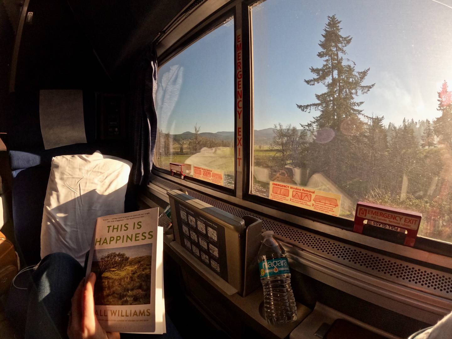 person reads a copy of the book This is Happiness next to a train window looking out onto a rural view