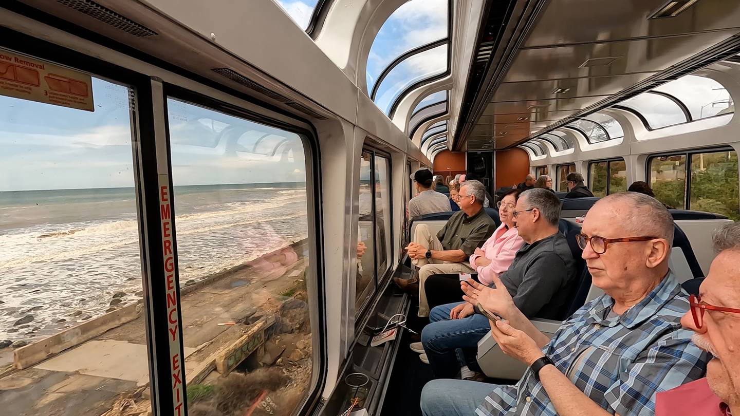 people seated in train car seats facing the window looking out toward the Pacific ocean