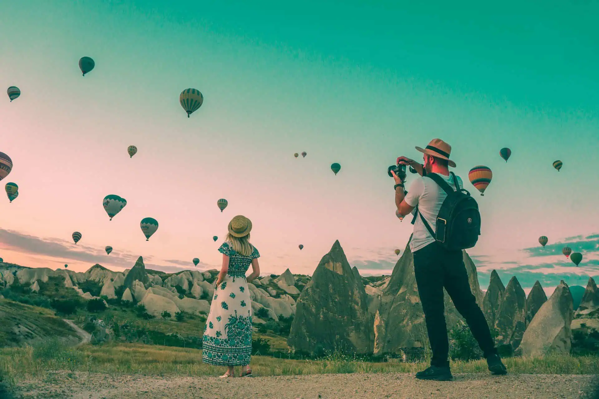 A man wearing a hat takes a photo of a woman in front of him also wearing a hat with her back to the camera. They are in Cappadocia, Turkey with hot air balloons and mountains in the distance.