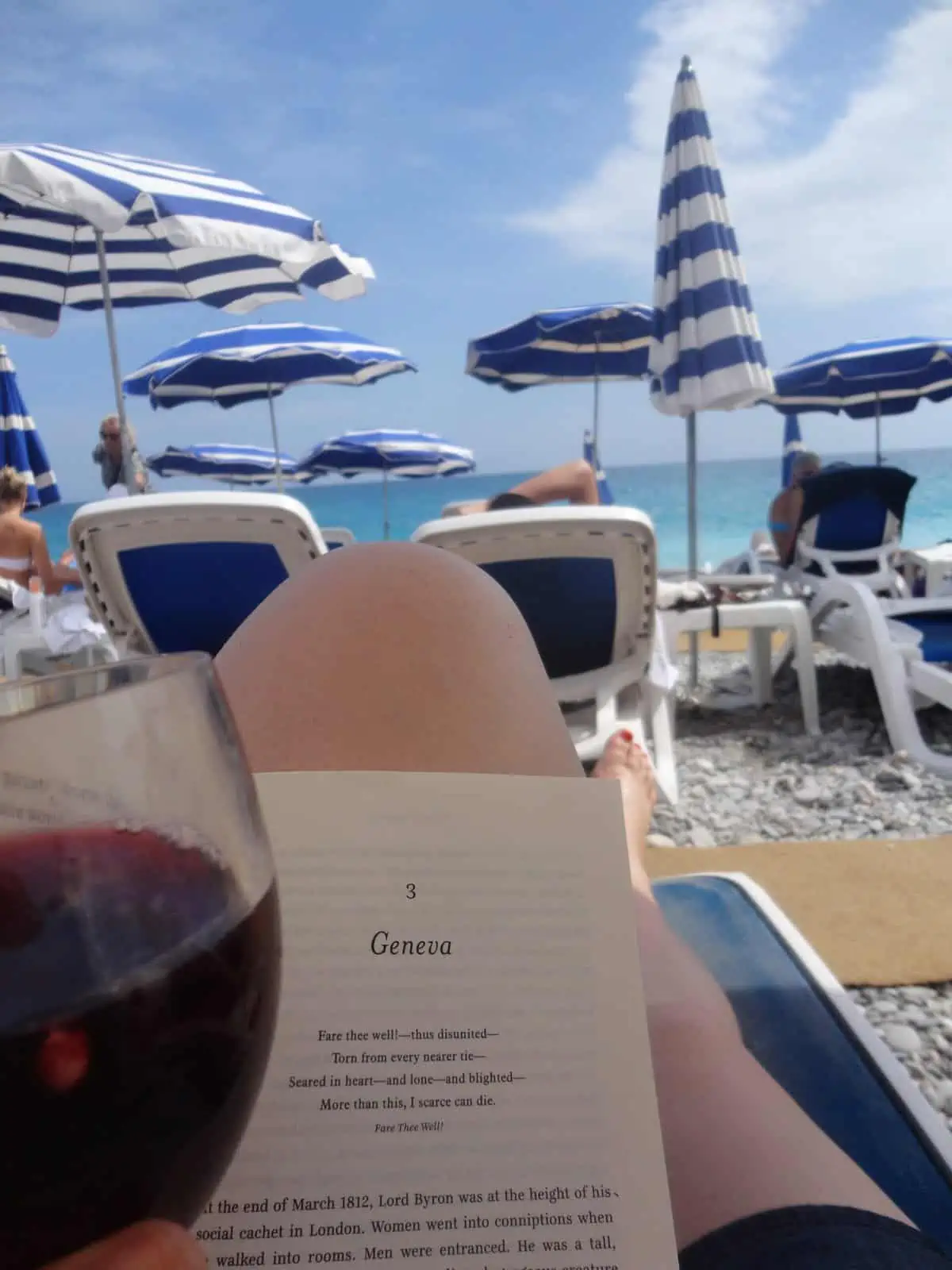 Shot of Riana's legs and holding a book and glass of sangria in front of them while sitting on a beach chair. There are rows of chairs and umbrellas in front of her on the beach.