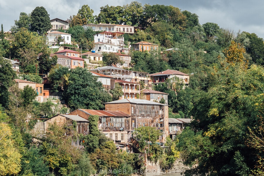 Old timber houses on the Rioni River in Kutaisi, surrounded by green foliage.