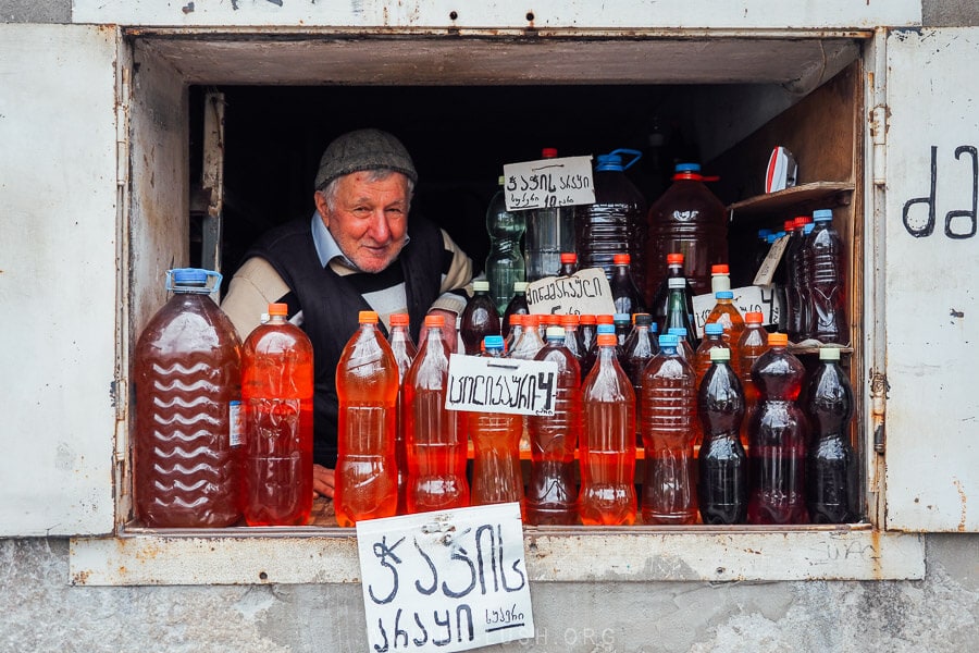 A man in a felted hat peers out between bottles of homemade liquor at the Green Bazaar market in Kutaisi, Georgia.