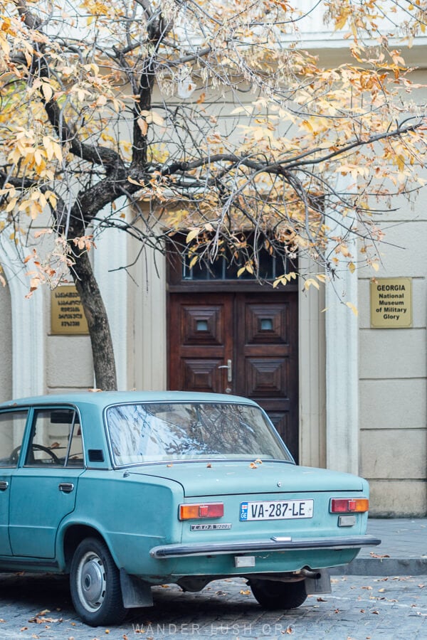 A vintage blue car parked under an autumn tree in Kutaisi, Georgia.