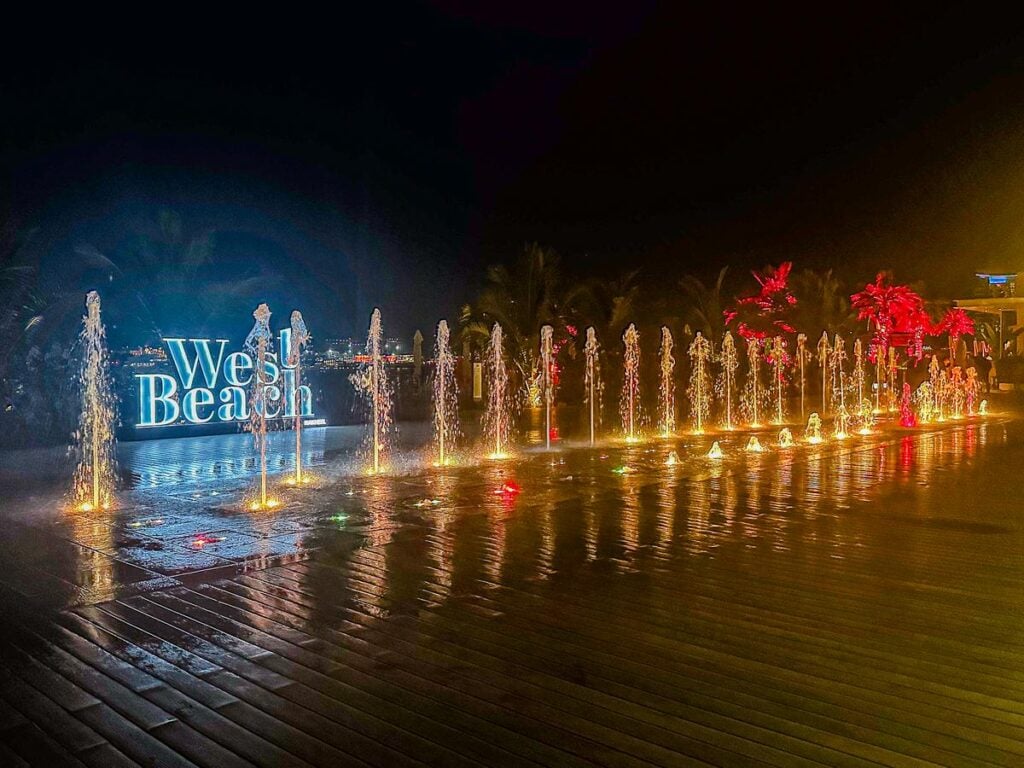 A nighttime view of lighted fountains and the illuminated 'West Beach' sign at Dubai's seaside promenade with tropical trees and wooden decking. 