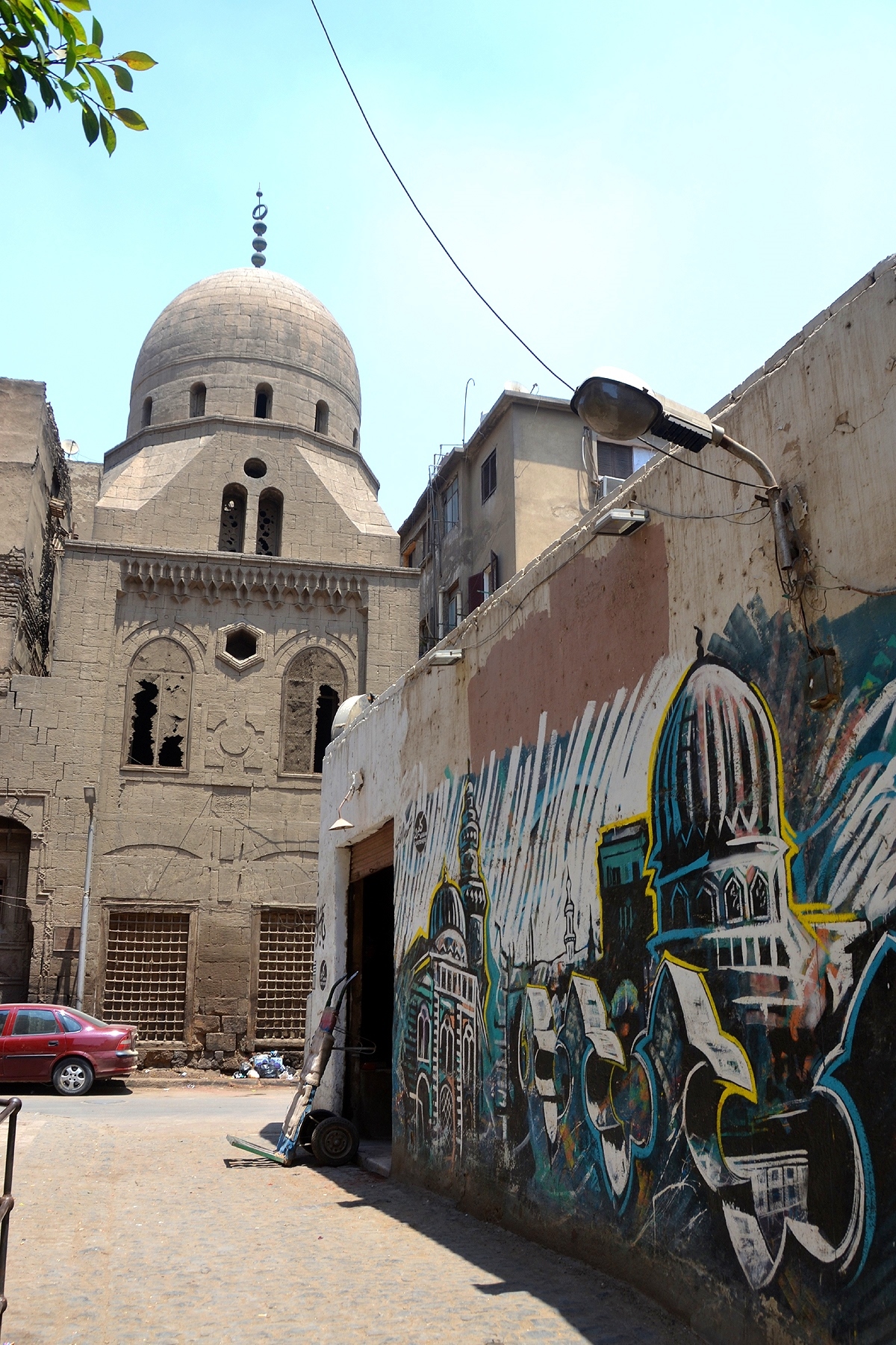 A street in Al Darb Al Ahmar shows a historic building topped with a dome and a mural of local historic buildings painted in bright blues and yellows. 