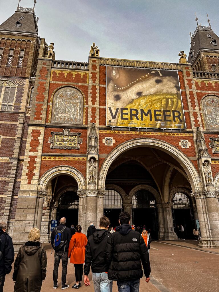 Tourists experiencing King's Day by walking under the Rijksmuseum archway featuring a Vermeer banner in Amsterdam, under a clear sky and surrounded by the museum's historic architecture.