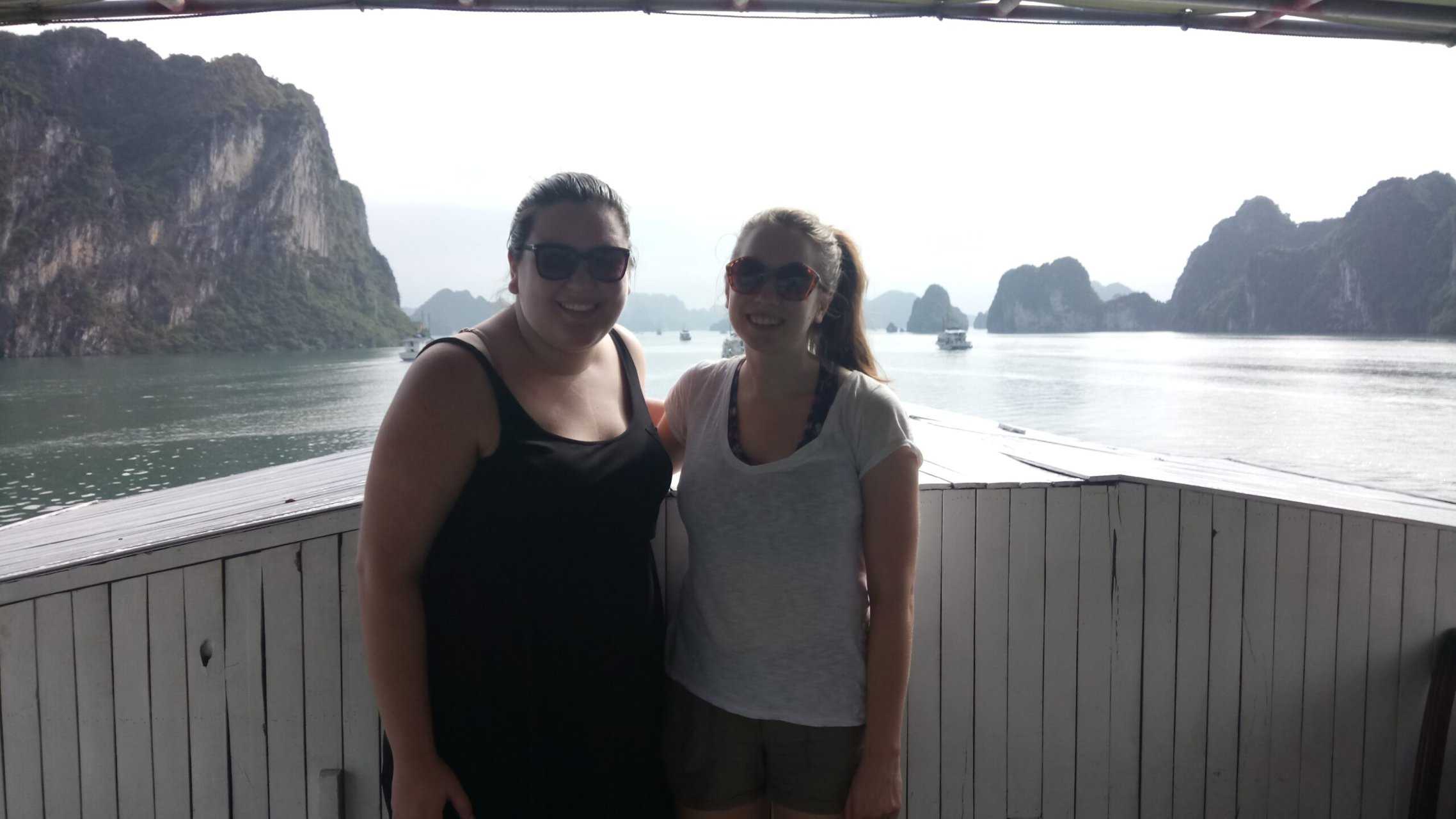 Riana and Emilie smiling on the back of their Halong Bay Cruise with limestone cliffs and water behind them