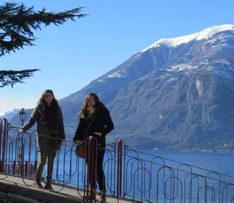 Riana and her friend Stephanie standing on a bridge over Lake Como with a mountain and water in the background