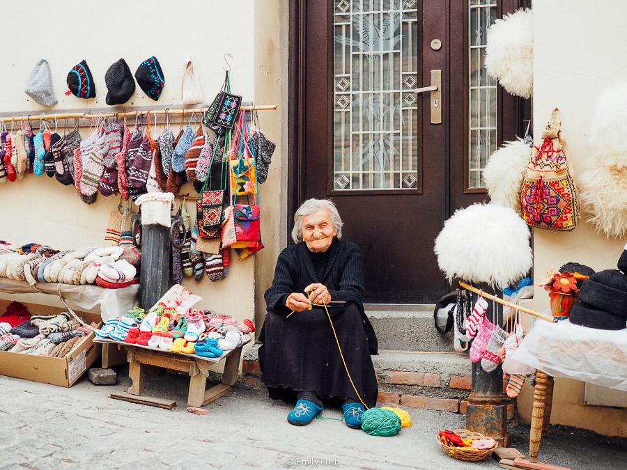 A woman in a black dress sits knitting on the street in Sighnaghi, Georgia.