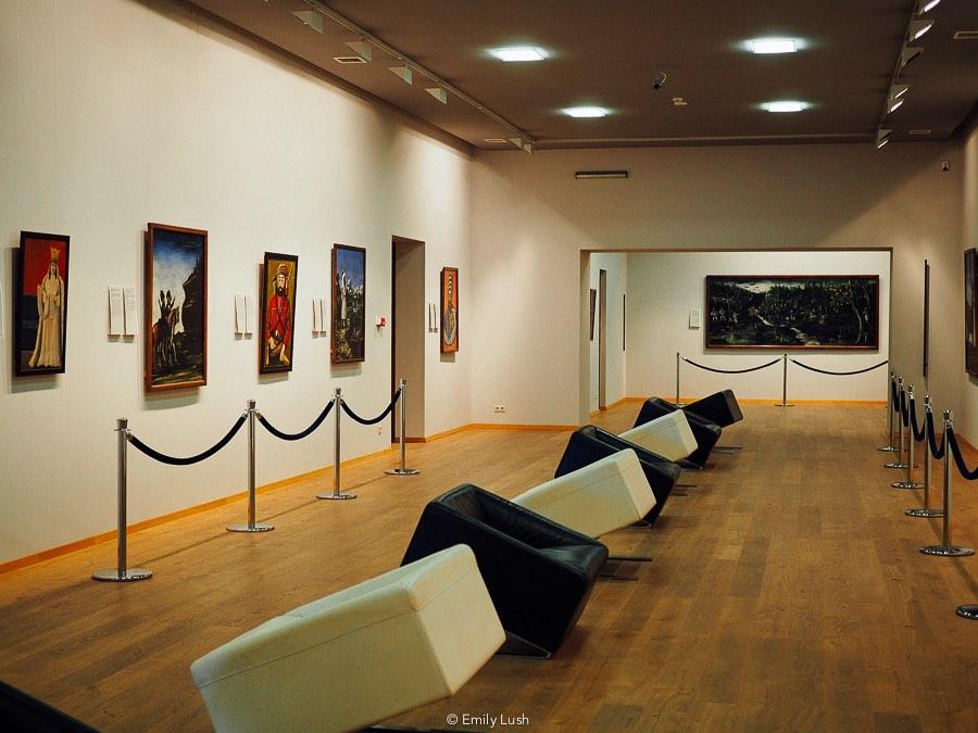 An art gallery with pictures on the wall, velvet ropes and a row of black and white chairs down the centre.