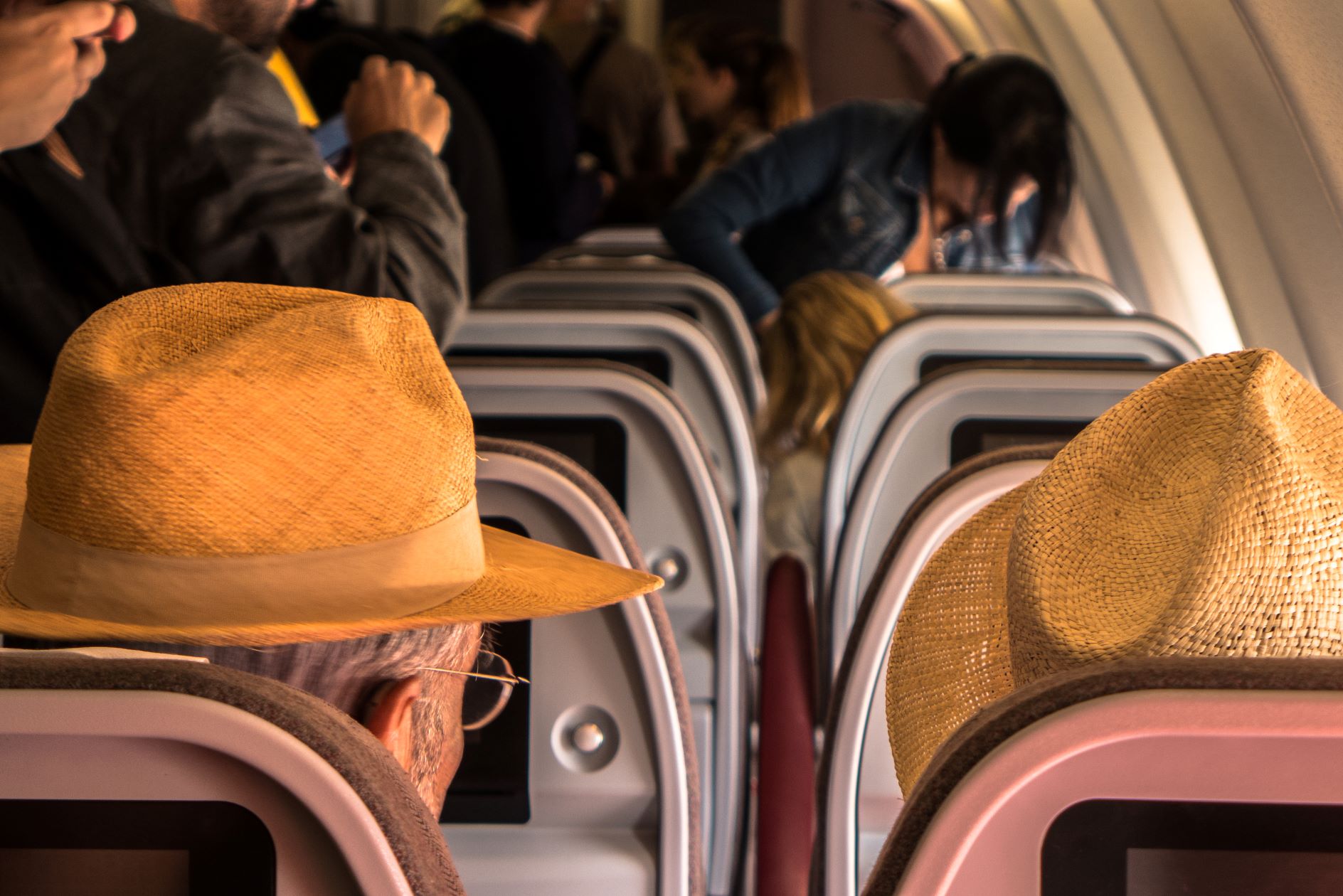 Two seniors wearing straw hats on an international flight among passengers waiting to deplane in another country.