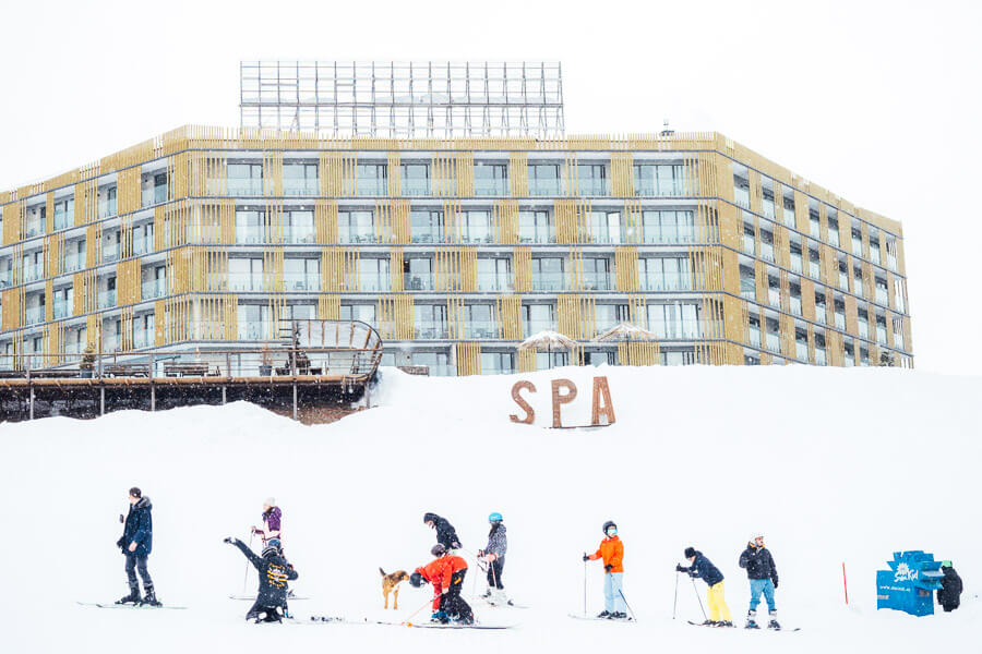 A line of skiers in colourful gear in front of a modern apart-hotel in Gudauri, Georgia.