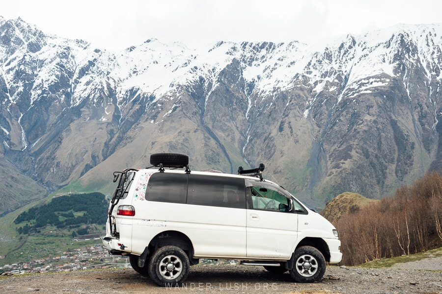 A white van parked in front of a snow-capped mountain in Kazbegi, Georgia.