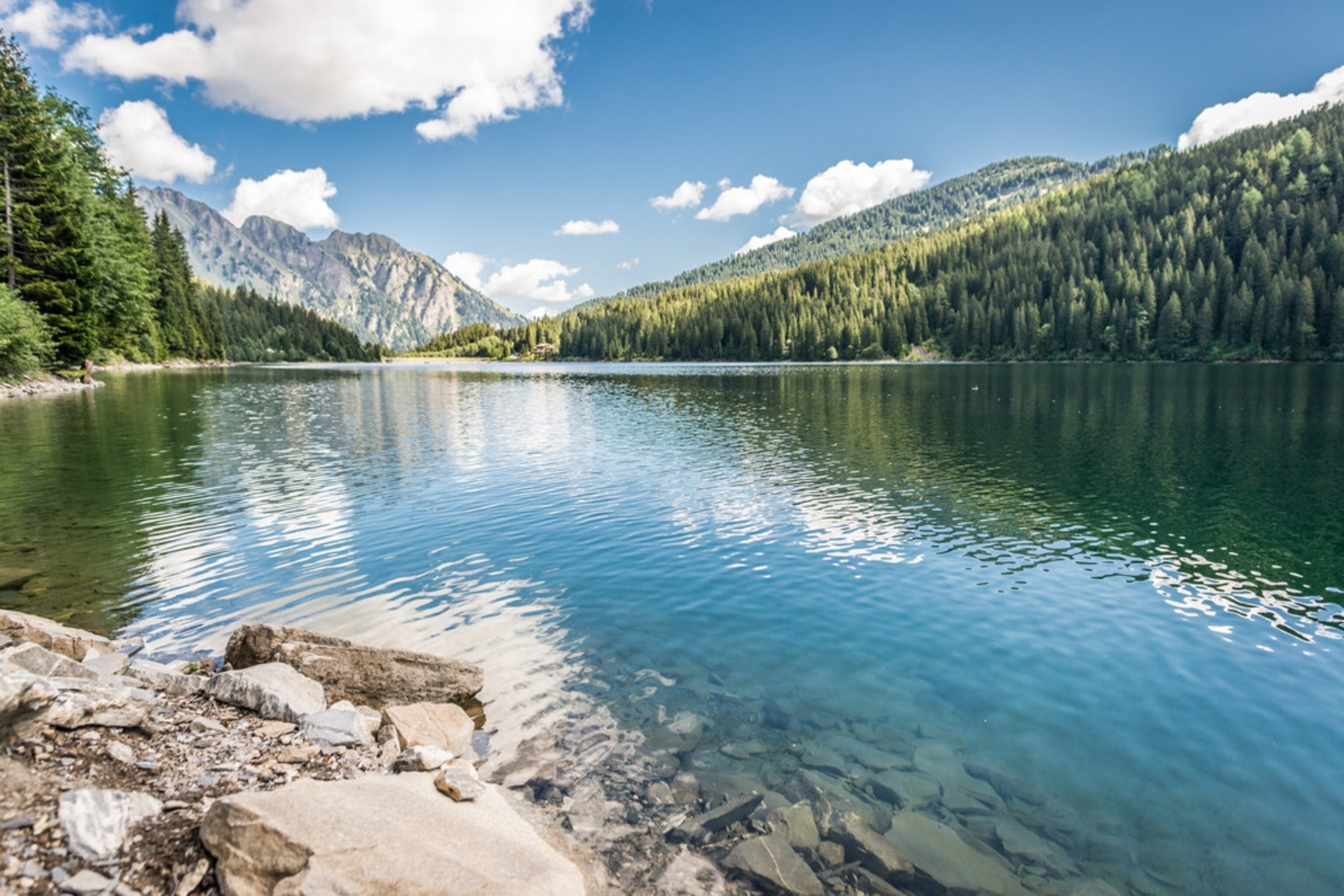 A bright blue alpine lake with forested slopes surrounding it and mountains behind.