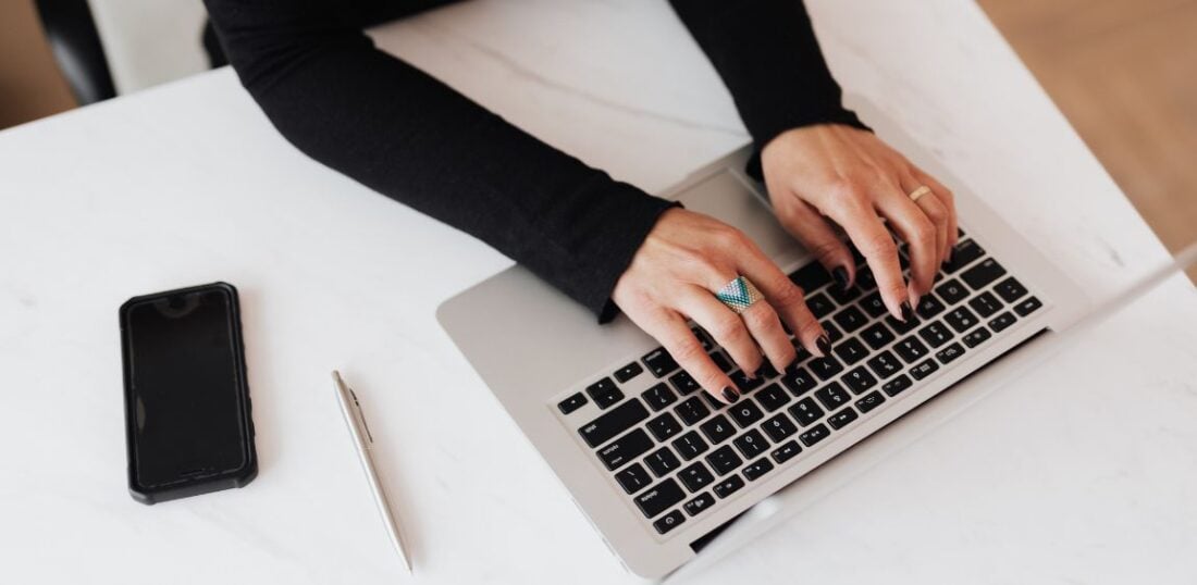 person typing on laptop on clear desk with phone and pen to the side 