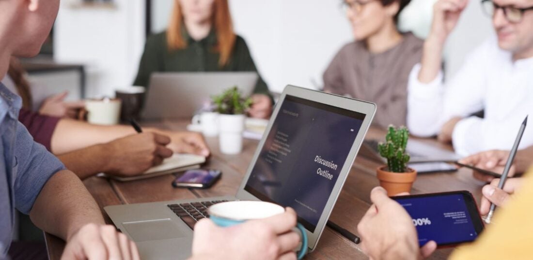 meeting room office space, multiple attendees sat with laptops and coffee. Mobile phones and small indoor plants on a shared desk