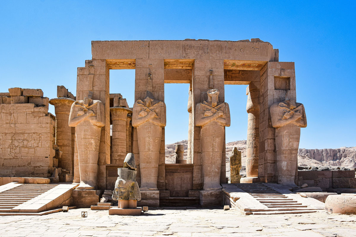 The Ramasseum in Luxor is shown with a row of columns and the fragmented head of a statue laying on the ground. 