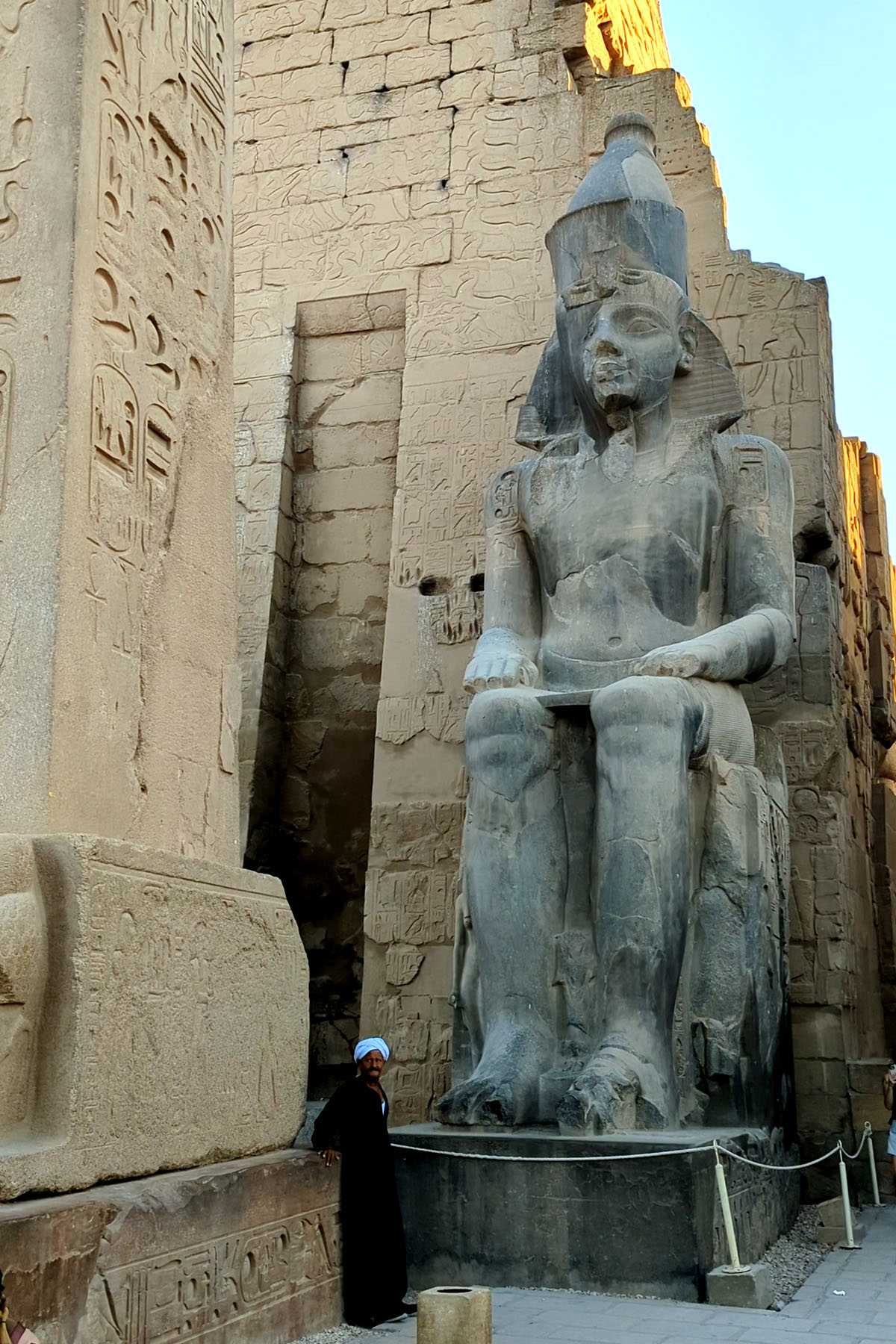 A statue of Ramses II stands outside the Luxor Temple in Egypt. An Upper Egyptian man stands at the base of the statue wearing a galabeya.