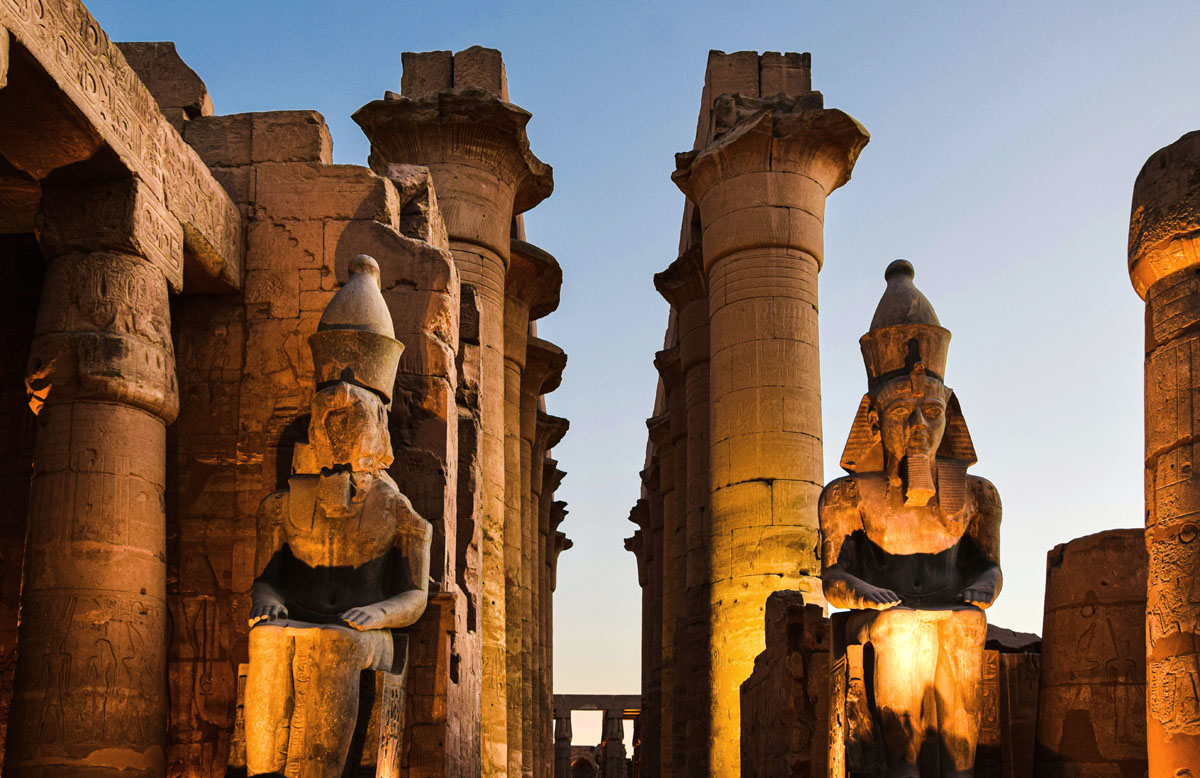 Rows of enormous columns and statues of Ramses II stand at Luxor Temple in the evening, lit up in a golden yellow light. 