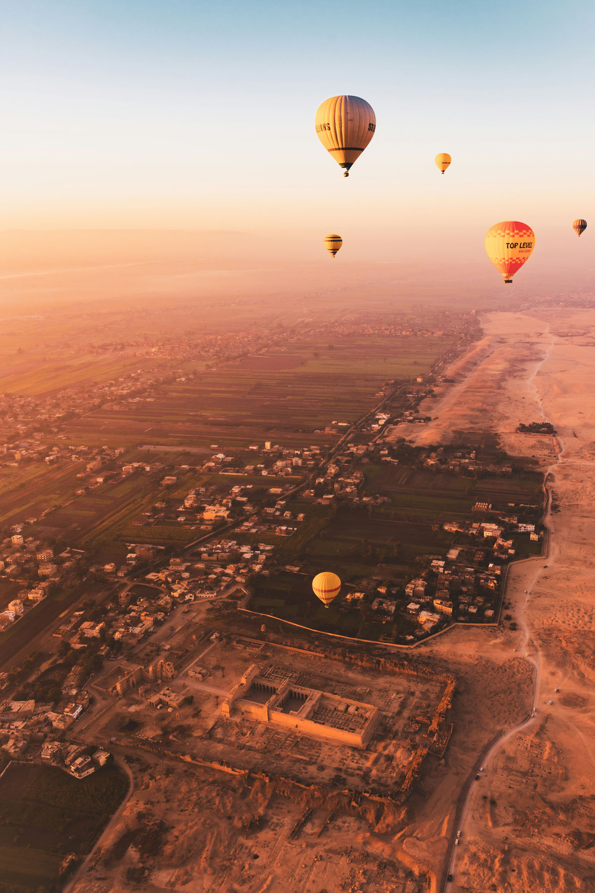 Hot air balloons fly over Luxor as the sun slowly rises. There is a pink and rosy glow.