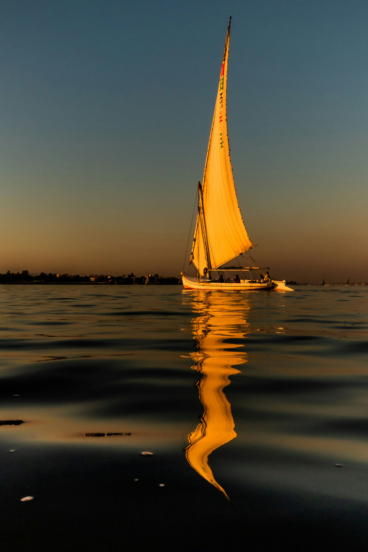 A felucca sails at sunset in Luxor, bathed in a golden light that is reflected in the water. 